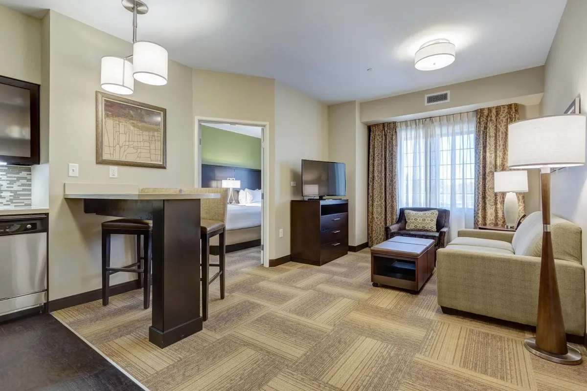 The image showcases a bright and inviting condo living area featuring a cozy sofa facing a large television, a modern bar table perfect for meals or conversation, and a window dressed with soft curtains letting in natural light. The setup suggests a perfect space for relaxing, socializing, and enjoying downtime with family or friends.