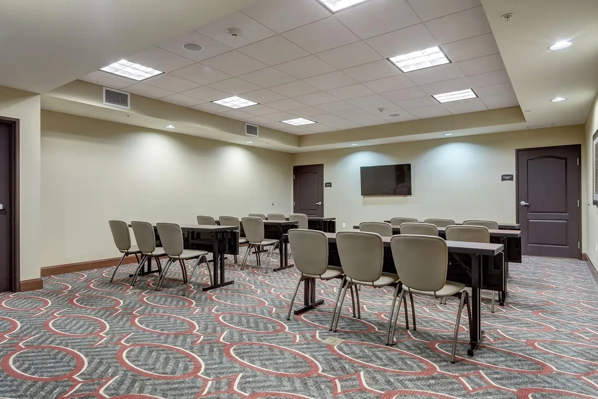A formal meeting room featuring tables and chairs arranged for group sessions, with a television mounted on the wall and visible doorways providing a sense of connectivity to other spaces. The setting is designed for gatherings, discussions, or collaborative activities, emphasizing functionality and comfort for small groups.