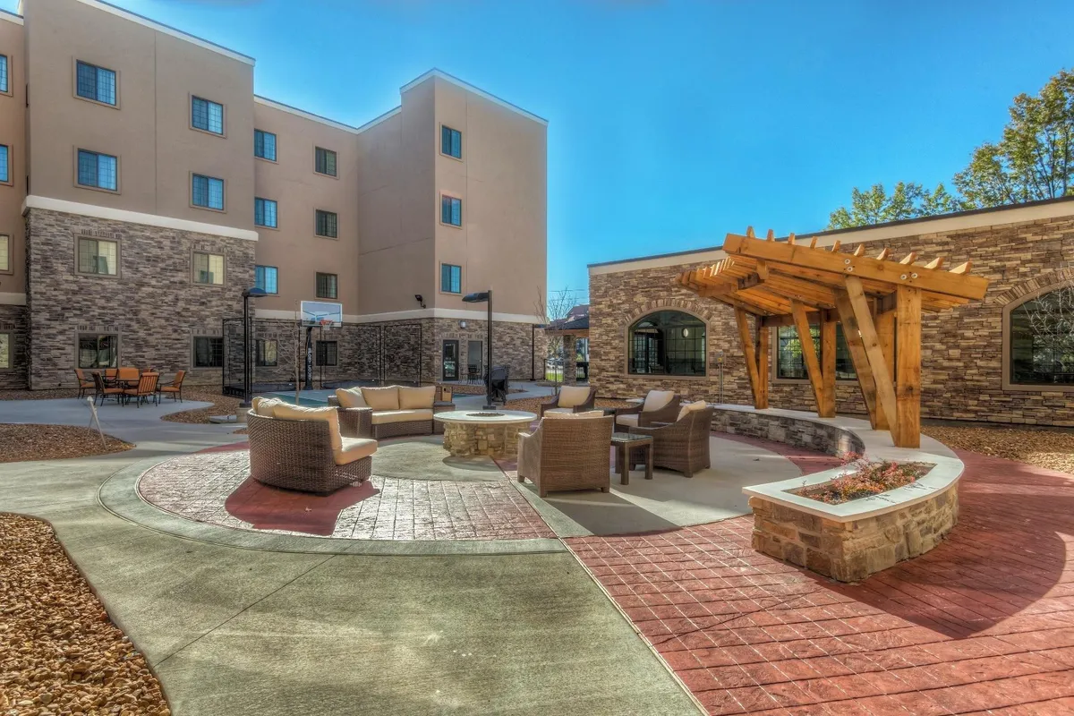 An outdoor communal area with comfortable couches, a stone fire pit, a wooden pergola, and the surrounding building in the background. The area invites guests to gather, relax, or spend time with friends and family, reflecting a sense of community and belonging.