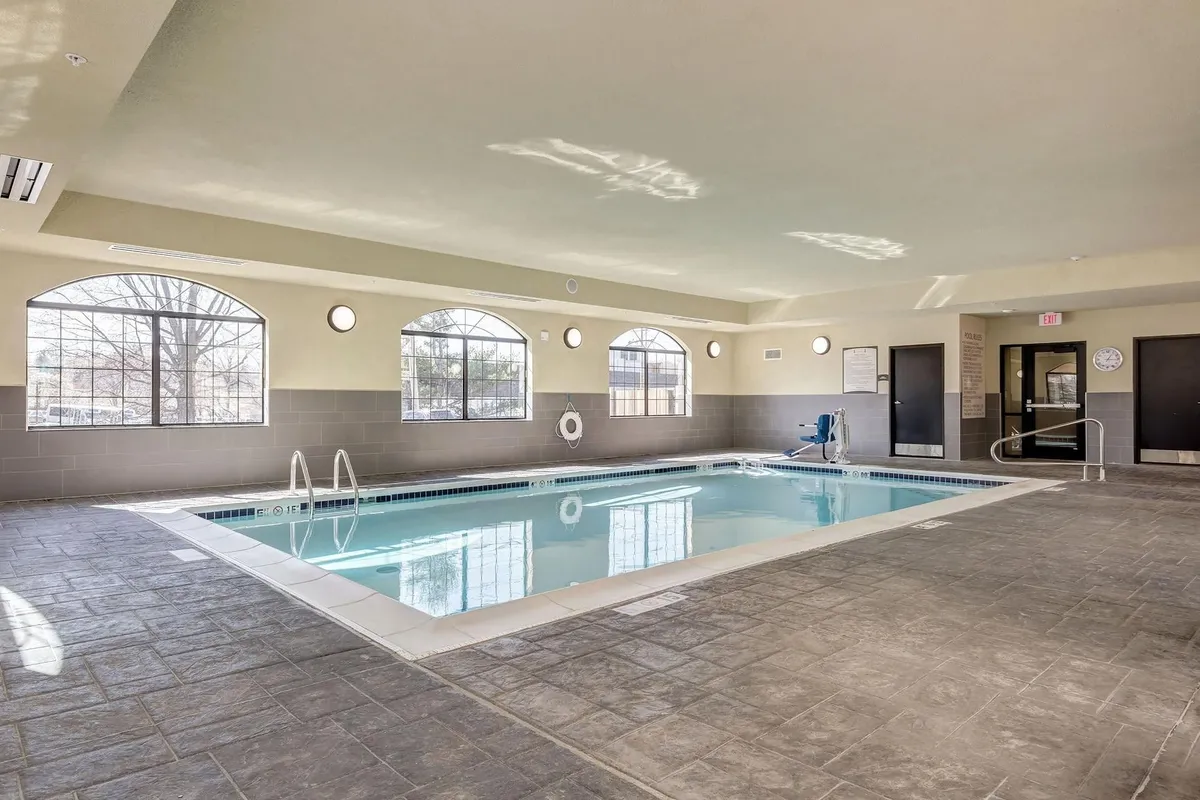 A spacious indoor swimming pool area, complete with large windows letting in natural light, poolside seating for families to relax, and visible safety equipment. The water is clear and inviting, while the seating accommodates both swimmers and those supervising children at play.