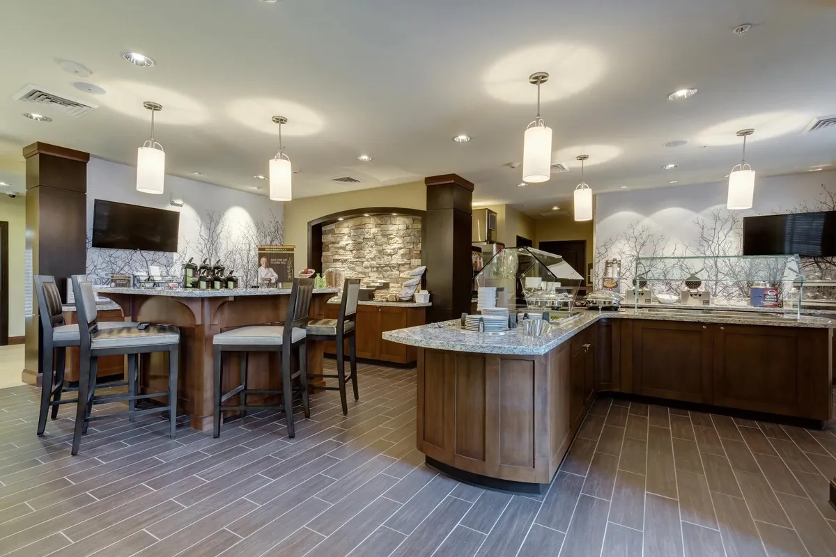 This image features a welcoming dining area with a granite countertop, wooden bar stools, and a stone wall feature next to the buffet space. The area looks inviting for socializing or enjoying a meal, with plenty of natural light and comfortable seating.