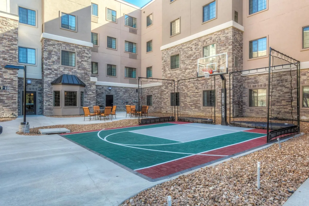 This exterior shot features a vibrant basketball court in the foreground, a welcoming seating area for spectators or family gatherings, and the modern facade of the Staybridge Suites building. The court is brightly lit by natural daylight, inviting energetic play and social interaction, with the building suggesting proximity to family accommodations.
