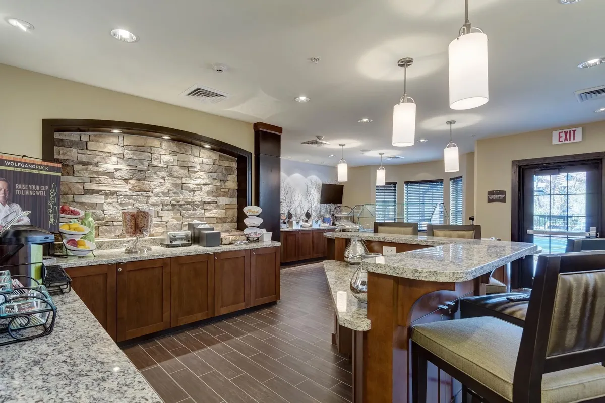 A warm, inviting interior common area featuring a granite countertop, stone wall, stylish lighting fixtures, and a food display. The scene suggests a comfortable spot for breakfast, coffee, or casual gatherings with other guests.